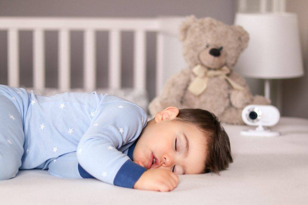 Cute little baby boy in light blue pajamas sleeping peacefully on bed at home with baby monitor