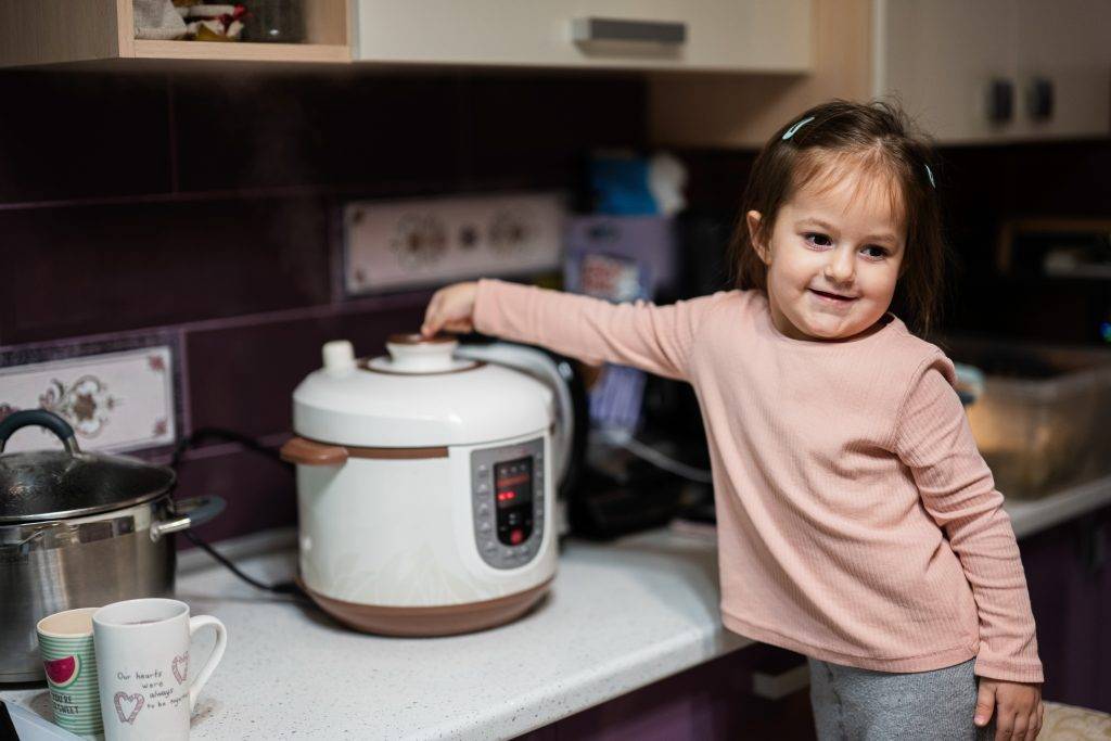 Baby girl prepare dinner at home kitchen with slow cooker and releases steam from the steamer.