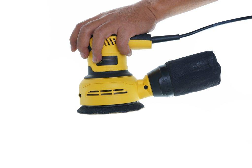 Closeup of a hand holding a yellow orbital sander on an isolated white background