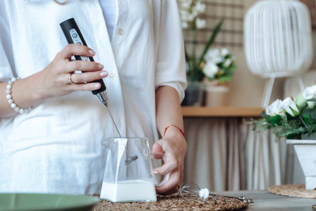 Mixing milk in glass pot by frother closeup. Female making foamy milk with handheld mixer. Housewife