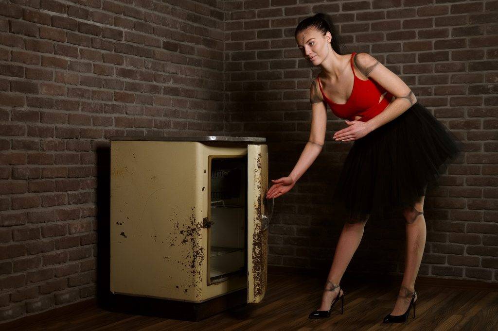 Sexy girl posing as a doll opening old broken dirty fridge.