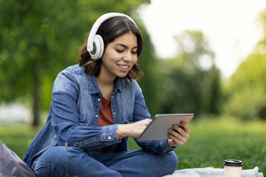 Young Middle Eastern Woman In Wireless Headphones Relaxing Outdoors With Digital Tablet