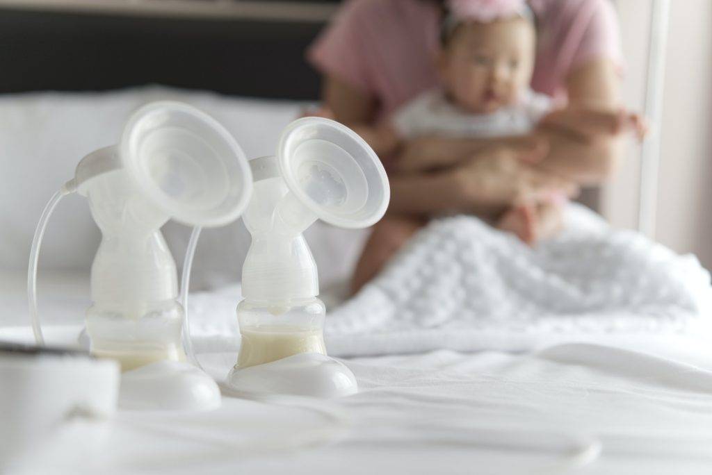 Breast milk pump's bottles and pump machine on the bed with mother hugging baby in background.