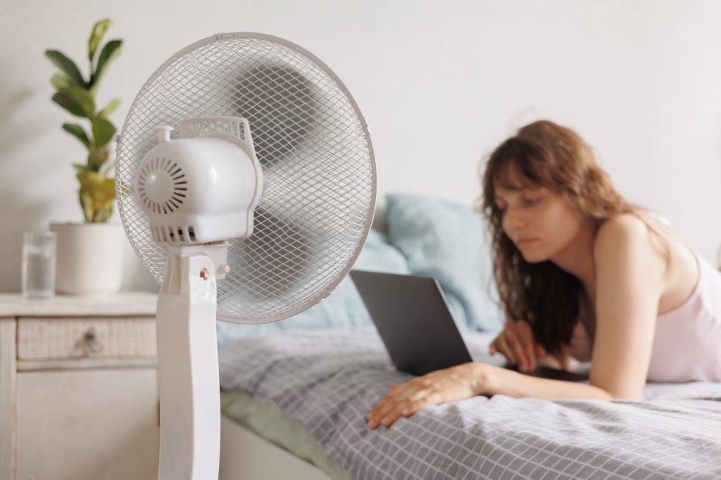 European woman cools down home with help of air conditioner fan, works with laptop in home office