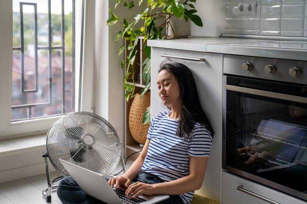 Relaxed Asian woman typing on laptop while cooling by floor fan at home.